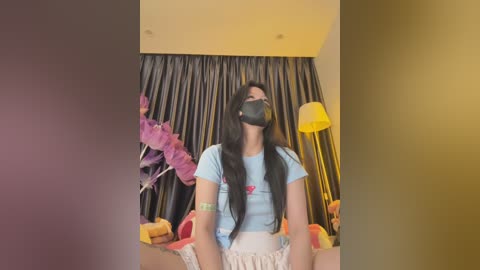 Media: Video of a young Asian woman with long black hair, wearing a face mask, light blue shirt, and white shorts, sitting on a bed with colorful pillows, surrounded by dark curtains and a yellow lamp.