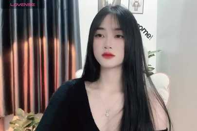 Media: Video of an East Asian woman with long black hair and fair skin, wearing a black V-neck top, in a modern room with curtains and potted plants.