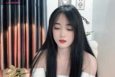 Media: Video of a young Asian woman with long black hair and pale skin, wearing a white strapless top, sitting in a room with a dark curtain and framed pictures on the wall.