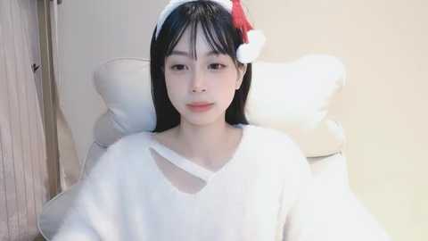 Media: A video of an Asian woman with long black hair, wearing a white sweater, a red Santa hat, and a neutral expression, seated on a white chair against a light beige wall.