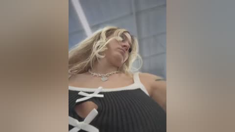 Media: Video of a blonde woman with wavy hair, wearing a black, ribbed tank top with white lace-up details and a silver necklace. Background shows a modern, industrial setting with gray walls and metallic structures.