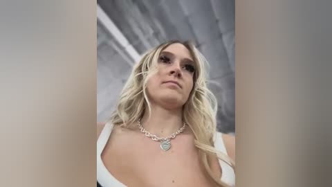 Media: Video of a blonde woman with wavy hair, wearing a white tank top and a silver heart necklace, looking upward in a dimly lit room with exposed concrete ceiling.