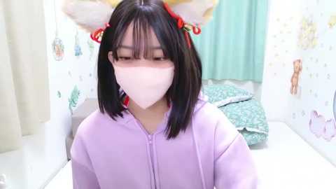 Media: A video of an East Asian woman with straight black hair and white face mask, wearing a light lavender hoodie, in a pastel-colored, cartoon-themed bedroom.