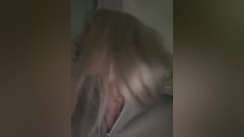 Media: Video of a blurred, blonde-haired woman with a partially open zipper on her light grey jacket, standing indoors with dim lighting.