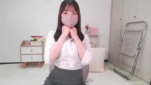 Media: Video of an East Asian woman with straight black hair, wearing a white blouse and gray skirt, adjusting a pink face mask in a minimalist, white-walled room with wooden furniture and a metal folding chair.