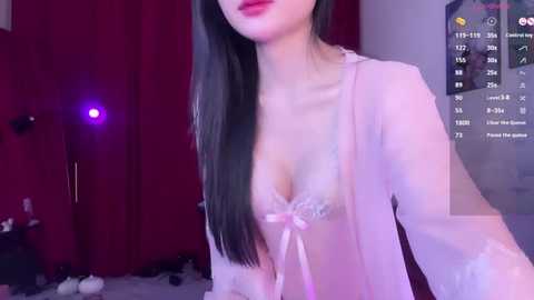 Media: Video of an East Asian woman with long black hair, wearing a sheer pink robe and revealing a lace bra, standing in a dimly lit room with red curtains and a digital clock.