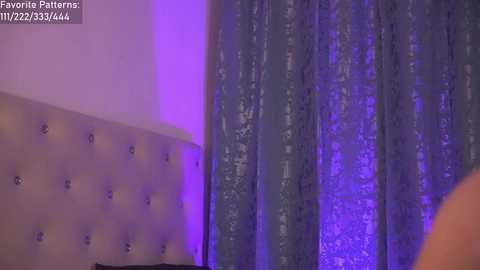 Media: A video of a plush, tufted headboard in a soft beige color against a backdrop of shimmering, purple sequined curtains. The image captures a luxurious, intimate bedroom setting with a subtle purple lighting effect.