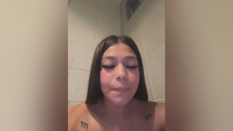 Media: A video of a young woman with long dark hair, closed eyes, and a neutral expression, standing in a tiled shower with visible nipples. She has small tattoos on her shoulders.