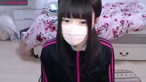 Media: Video of an Asian woman with long black hair, wearing a face mask, black jacket, and floral-patterned bedspread in a cluttered, brightly lit bedroom.