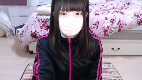 Media: A video of an Asian woman with long black hair, wearing a face mask, black jacket with pink stripes, and seated on a striped rug in a bedroom with a floral bedspread.