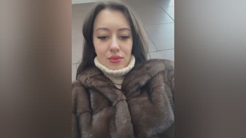 Media: Video of a young woman with fair skin and long brown hair, wearing a brown fur coat and white turtleneck sweater, indoors against a tiled floor.