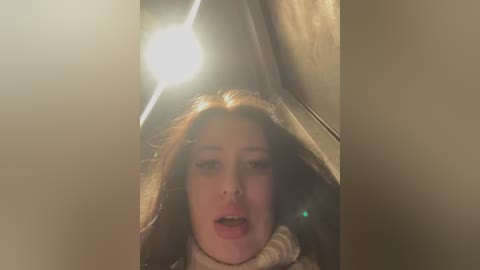 Media: A blurry video of a young woman with light skin and long, wavy brown hair, her mouth open, taken inside a dimly lit car, with a bright light source creating a halo effect.