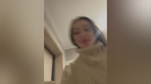 Media: Blurry video of a woman with light skin, brown hair, and wearing a beige sweater, standing in a narrow hallway with beige walls and a textured ceiling.