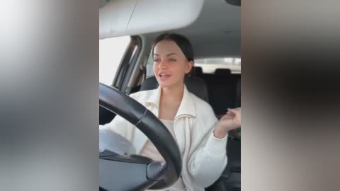 Media: Video of a young woman with light skin and dark hair, wearing a white jacket, smiling, driving a car with a black steering wheel and grey seats.