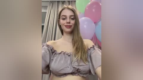 Video of a smiling, fair-skinned, blonde woman with long hair, wearing a light lavender off-shoulder top, standing in a room with a pink and blue balloon backdrop.