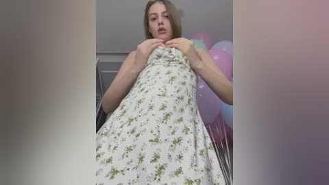 Video of a young woman with fair skin and light brown hair, wearing a white floral dress, holding balloons in a room with soft lighting.