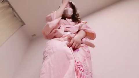 Media: A video of a person in a pink robe with floral patterns, sitting on the floor, wearing a surgical mask, and covering their face with one hand, in a dimly lit room.