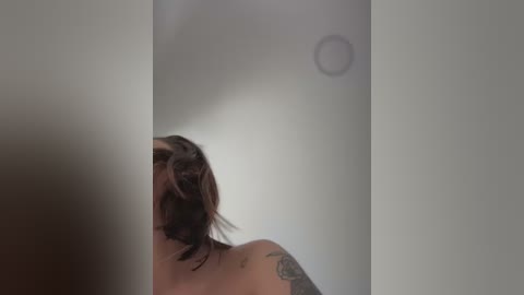 Media: Video of a woman with short, dark hair, wearing a black bra, leaning against a white wall, with a visible tattoo on her left shoulder.