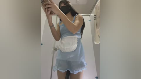 Media: Video of a slender East Asian woman with long black hair in a blue, frilly maid dress, holding a phone in a narrow, white-walled bathroom.