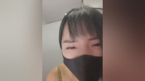 Media: Video of an Asian woman with short black hair, wearing a black mask, in a dimly lit room, her eyes partially visible, looking distressed.