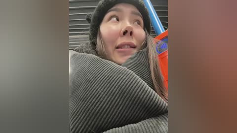 Media: Video of an Asian woman with long hair, wearing a black hat and gray ribbed sweater, partially obscured by a blue and orange drink cup, standing in a subway car.