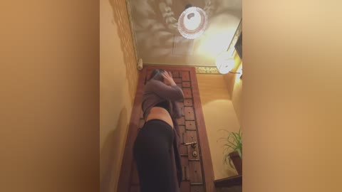 Media: Video of a woman in a tight black dress, bending over a wooden chest, in a narrow hallway with a circular ceiling light and a potted plant.