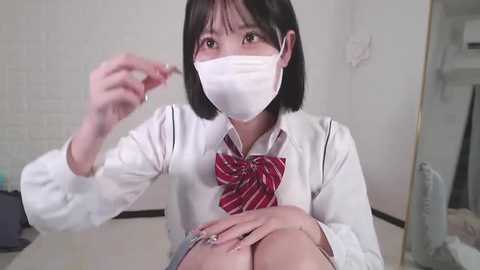 Media: A video of an Asian woman with short black hair, wearing a white face mask, red bow tie, white blouse, and blue shorts, sitting on a floor. She holds a cigarette in her right hand.