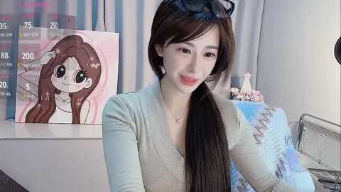 Media: Video of a smiling young Asian woman with long brown hair, wearing a light gray top, seated in a cozy room with a framed anime character poster and a fluffy pillow.