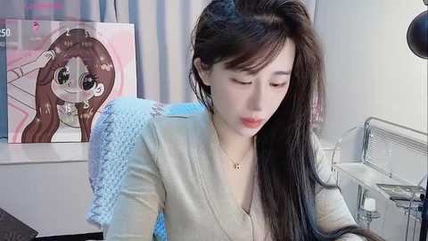 Media: A video of an Asian woman with long black hair, wearing a beige cardigan, sitting at a desk. A colorful anime poster hangs on the wall behind her.