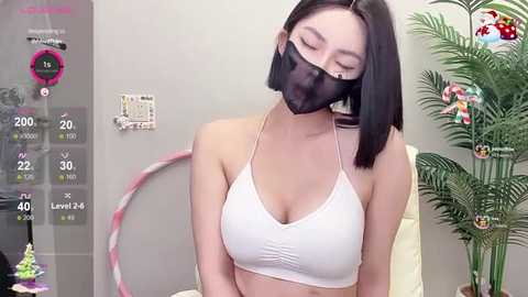 Media: A video of an East Asian woman with straight black hair, wearing a white sports bra and a black face mask, standing in a room with a potted plant and a whiteboard.