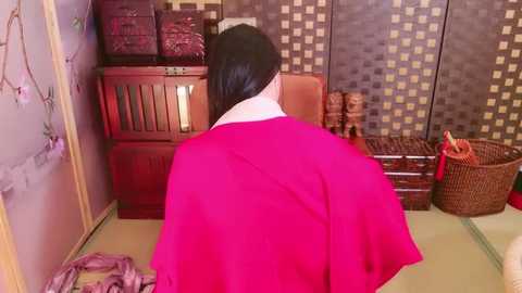 Media: Video of a woman with long black hair wearing a bright pink kimono, standing in a traditional Japanese room with wooden furniture, woven baskets, and a tatami mat floor.