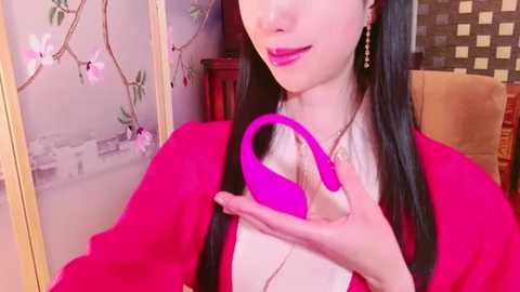 Media: Video of an Asian woman with long black hair, wearing a red cardigan and a white top, holding a bright pink vibrator. Background features a traditional Japanese screen and a wooden chair.