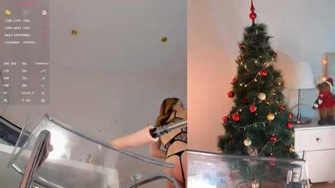 Media: Video of a woman in a striped top, partially visible, adjusting a Christmas tree in a modern living room with white walls, glass furniture, and a white table.