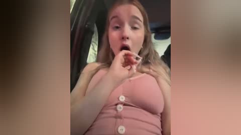 Media: Video of a young Caucasian woman with fair skin and long brown hair, wearing a pink ribbed tank top with large buttons. She is licking a lollipop, eyes closed in pleasure, inside a car.