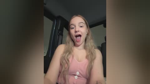 Media: Video of a young, fair-skinned woman with long, wavy blonde hair, wearing a pink top, sticking out her tongue while sitting in a dimly lit room with dark furniture.