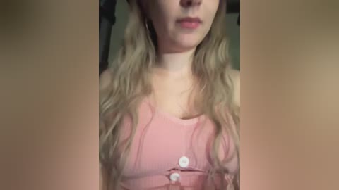 Media: Video of a young woman with long, wavy blonde hair, wearing a pink tank top with a single button. The background is blurred, possibly indoors with muted lighting.