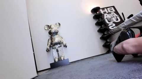 Video of a person squatting with a dumbbell, wearing a gray shirt and black shoes, standing beside a metallic Mickey Mouse statue and a black weight rack against a white wall.