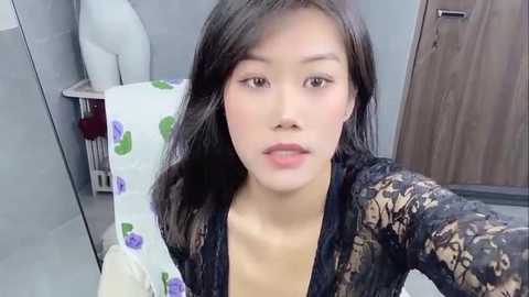 Media: Video of a young Asian woman with long black hair, fair skin, and a medium build, wearing a black lace top. She sits in a bathroom with a white towel and purple-patterned toilet roll visible.