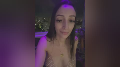 Media: A video of a young woman with long dark hair and fair skin, wearing a low-cut, sparkly top, standing outside at night with a city skyline in the background.