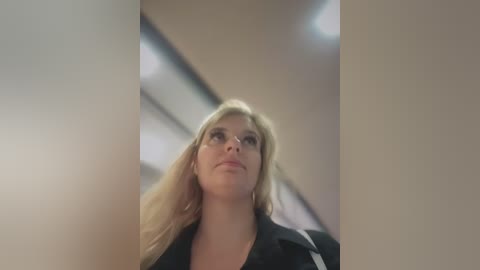 Media: Video of a blonde woman with long hair, wearing a black blazer, looking up intently, in a modern, minimalist indoor setting with beige walls and recessed lighting.