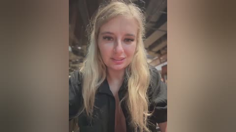 Media: Video of a young, fair-skinned woman with long, wavy blonde hair, wearing a black shirt. She has a slight smile, and the background shows an industrial setting with pipes and machinery.