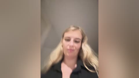 Media: A blurred video of a fair-skinned, blonde woman with long hair, wearing a black jacket, standing in a dimly lit, beige hallway.