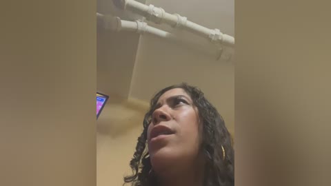 Media: Video of a young woman with curly dark hair and gold hoop earrings, looking upward at a fluorescent light fixture with pipes.