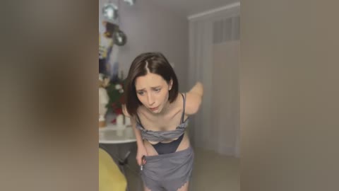 Media: Video of a slender Caucasian woman with short brown hair, wearing a grey bra and panties, bending over in a dimly lit room. Background features a yellow chair and a decorative plant.