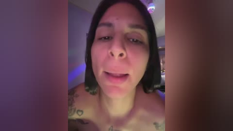 A video of a topless woman with shoulder-length black hair, tattoos on her shoulders, and a slight smile, taken in a dimly lit room with purple lighting.