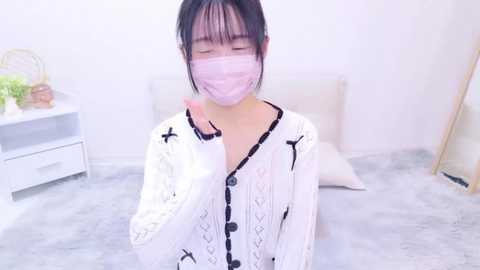 Media: Video of an Asian woman with straight black hair, wearing a white lace cardigan, pink mask, and light pink shirt, sitting on a gray carpet in a minimalist white room.