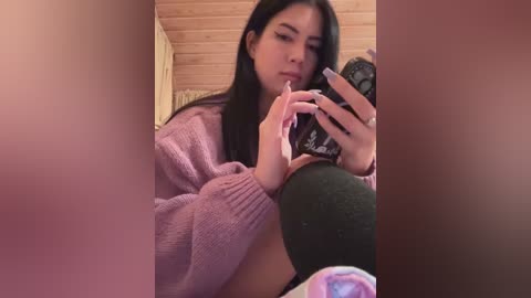 Media: A video of a young woman with long black hair, wearing a pink sweater, holding a phone, sitting on a wooden bench in a cozy room.