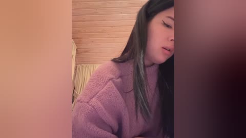 Media: Video of a young Asian woman with long black hair, wearing a pink sweater, leaning against a wooden wall with a radiator behind her.