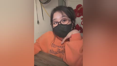 Media: Video of an Asian woman with black hair in pigtails, wearing glasses, a black face mask, and an orange University of Massachusetts sweatshirt. Background features red flowers and a fan.