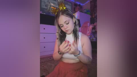 Media: Video of a young, fair-skinned woman with long, wavy, blonde hair, wearing a white crop top and red skirt, kneeling on a carpeted floor, examining her breasts.